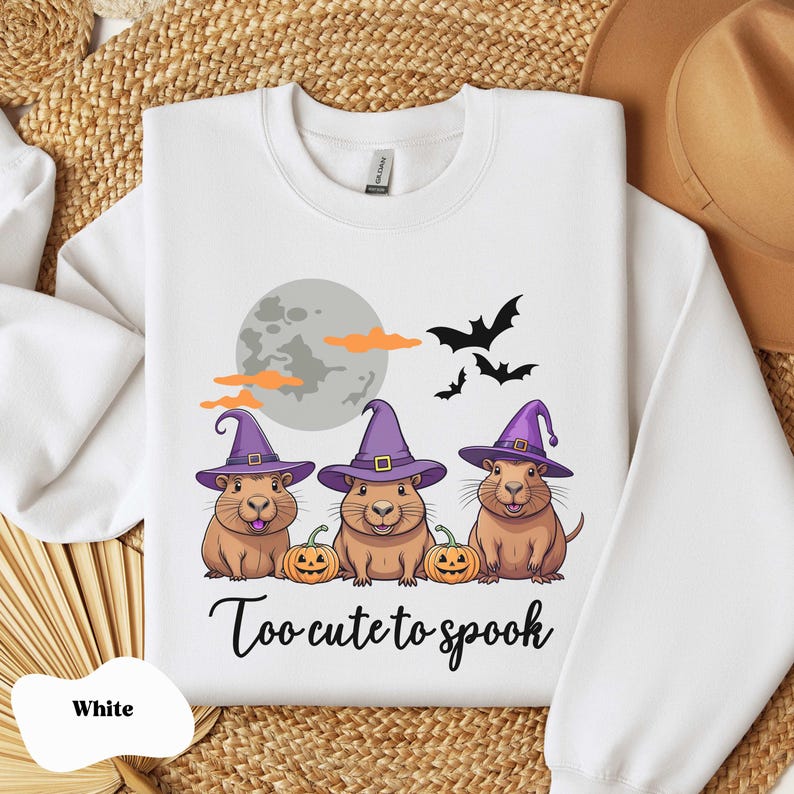 Op de afbeelding: Wit sweatshirt met een afbeelding van drie cartoon capybara's die toverhoeden dragen en pompoenen vasthouden. De tekst "Too cute to spook" is onder de capybara's gedrukt.