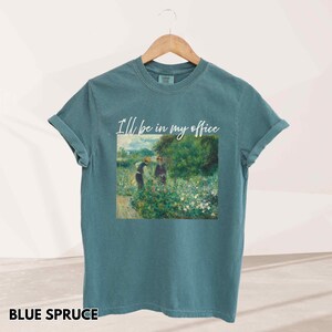 May include: Blue spruce t-shirt with rolled sleeves. The shirt features a painting of two people in a garden with the text "I'll be in my office" in white script. The shirt is hanging on a wooden hanger.