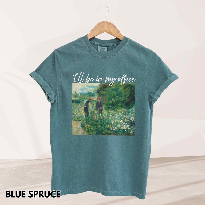 May include: Blue spruce t-shirt with rolled sleeves. The shirt features a painting of two people in a garden with the text "I'll be in my office" in white script. The shirt is hanging on a wooden hanger.
