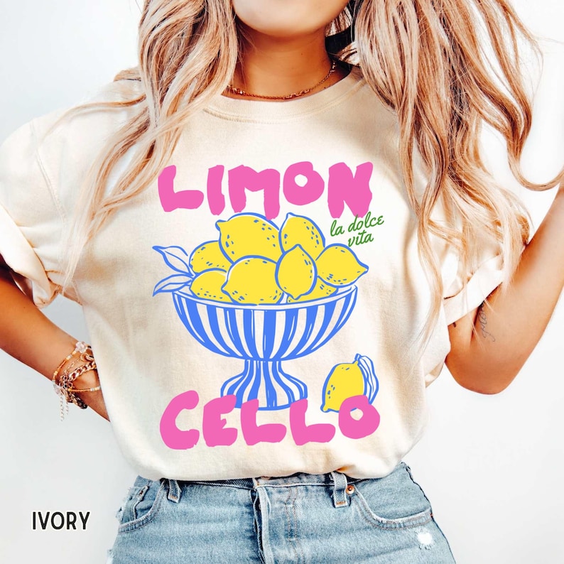 May include: Ivory-colored t-shirt with the words "LIMON CELLO" in pink, and a bowl of yellow lemons with blue stripes. The phrase "la dolce vita" is in green. A fun, summery design.