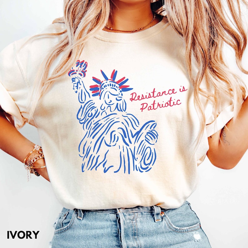 Pu&ograve; includere: T-shirt color avorio con un'illustrazione blu e rossa della Statua della Libert&agrave;. Il testo "Resistance is Patriotic" &egrave; stampato in rosso. La maglietta &egrave; in stile casual.