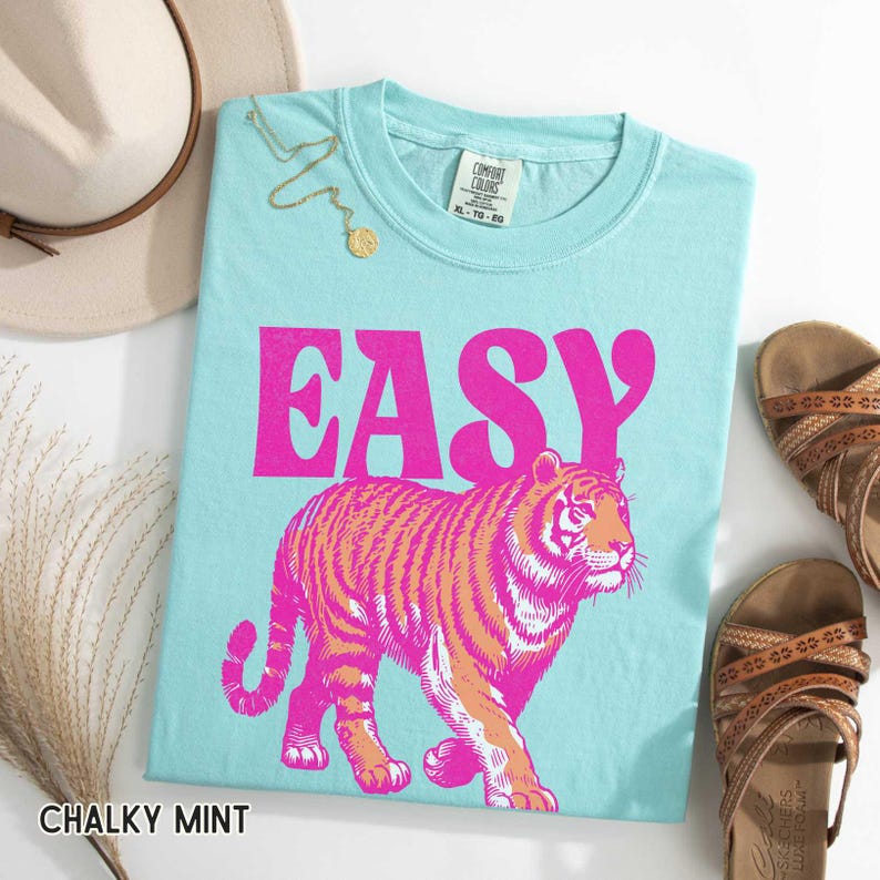 K&ouml;nnte beinhalten: Ein hellblaues T-Shirt mit dem Wort "EASY" in leuchtend rosa Buchstaben und einer rosa-orangenen Tigergrafik. Das Shirt liegt neben einem beigen Hut, einer goldenen Halskette und braunen Sandalen. Das Shirt ist mit "Chalky Mint" beschriftet.