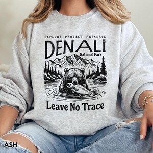 May include: Gray sweatshirt with a black and white graphic design of a bear holding a fish. The graphic includes the text "Explore Protect Preserve Denali National Park Leave No Trace".