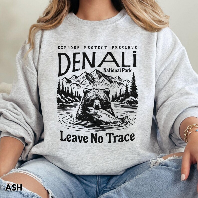 May include: Gray sweatshirt with a black and white graphic design of a bear holding a fish. The graphic includes the text "Explore Protect Preserve Denali National Park Leave No Trace".
