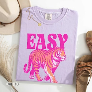 K&ouml;nnte beinhalten: Orchideenfarbenes T-Shirt mit dem Wort "EASY" in leuchtendem Pink und einer Tigerillustration in Pink und Orange. Das Shirt ist Teil einer Casual-Wear-Kollektion. Eine goldene Halskette und Sandalen sind ebenfalls zu sehen.