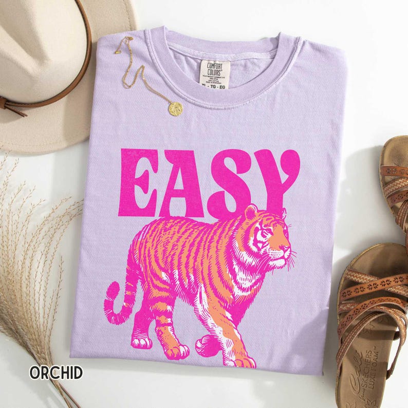 K&ouml;nnte beinhalten: Orchideenfarbenes T-Shirt mit dem Wort "EASY" in leuchtendem Pink und einer Tigerillustration in Pink und Orange. Das Shirt ist Teil einer Casual-Wear-Kollektion. Eine goldene Halskette und Sandalen sind ebenfalls zu sehen.