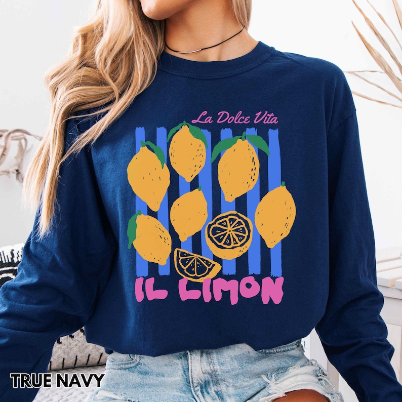 May include: A navy blue long-sleeve sweatshirt featuring a lemon graphic with the text "La Dolce Vita" and "Il Limon". The design includes yellow lemons and blue vertical stripes.