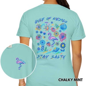 May include: A light blue t-shirt with a colorful graphic design on the back. The graphic features a flamingo, flowers, a seashell, sunglasses, and the text "Gulf of Mexico" and "Stay Salty". The front of the shirt has a small flamingo graphic with the text "Stay Salty".