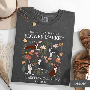 May include: A dark gray t-shirt with a white graphic design featuring Boston Terriers in a flower market setting. The text reads "The Boston Terrier Flower Market, Los Angeles, California Est 2025".