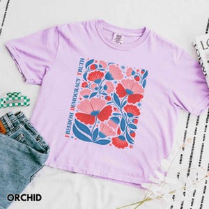 May include: Orchid-colored t-shirt with a floral design in red, pink, and blue. The words "FREEDOM DEMOCRACY TRUTH" are printed vertically on the left side of the floral design. The word "ORCHID" is printed at the bottom.