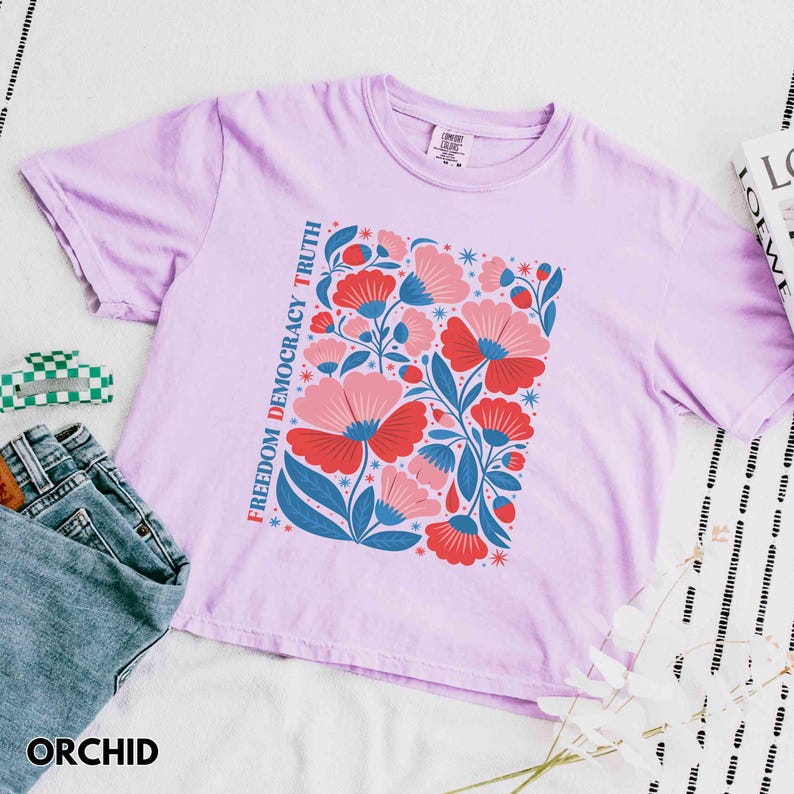 May include: Orchid-colored t-shirt with a floral design in red, pink, and blue. The words "FREEDOM DEMOCRACY TRUTH" are printed vertically on the left side of the floral design. The word "ORCHID" is printed at the bottom.