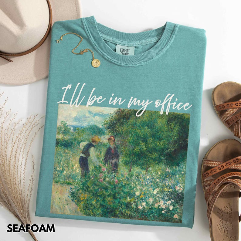 May include: Seafoam green t-shirt with the text "I'll be in my office" above a painting of two people in a garden. A gold necklace, brown sandals, and a beige hat are also visible.