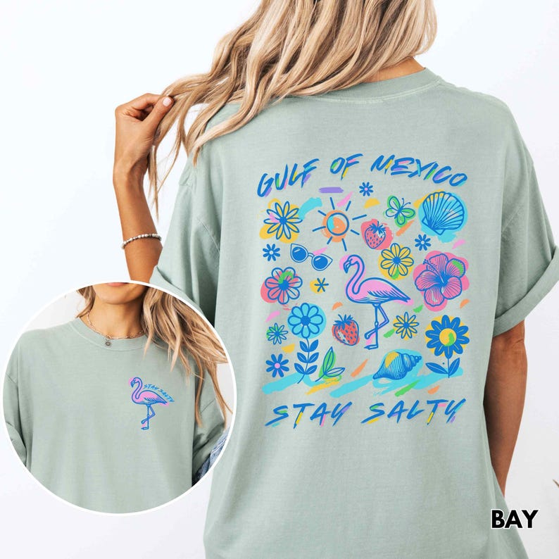 May include: A light blue t-shirt with a graphic print of a pink flamingo, flowers, a seashell, sunglasses, a sun, and strawberries. The text "GULF OF MEXICO" and "STAY SALTY" is printed on the shirt.