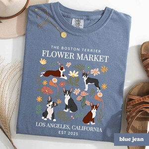 May include: A blue t-shirt with a floral design and Boston Terrier dogs. The text "The Boston Terrier Flower Market" is printed on the shirt, along with "Los Angeles, California" and "Est 2025".