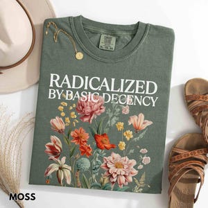 May include: Moss green t-shirt with the text "RADICALIZED BY BASIC DECENCY" and a floral design. A gold necklace, a beige hat, and brown sandals are also visible.