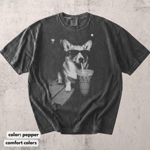 May include: Pepper-colored t-shirt featuring a graphic of a corgi wearing sunglasses and drinking from a to-go cup with a straw. The dog is sitting on a sidewalk. The shirt has a vintage, worn look.