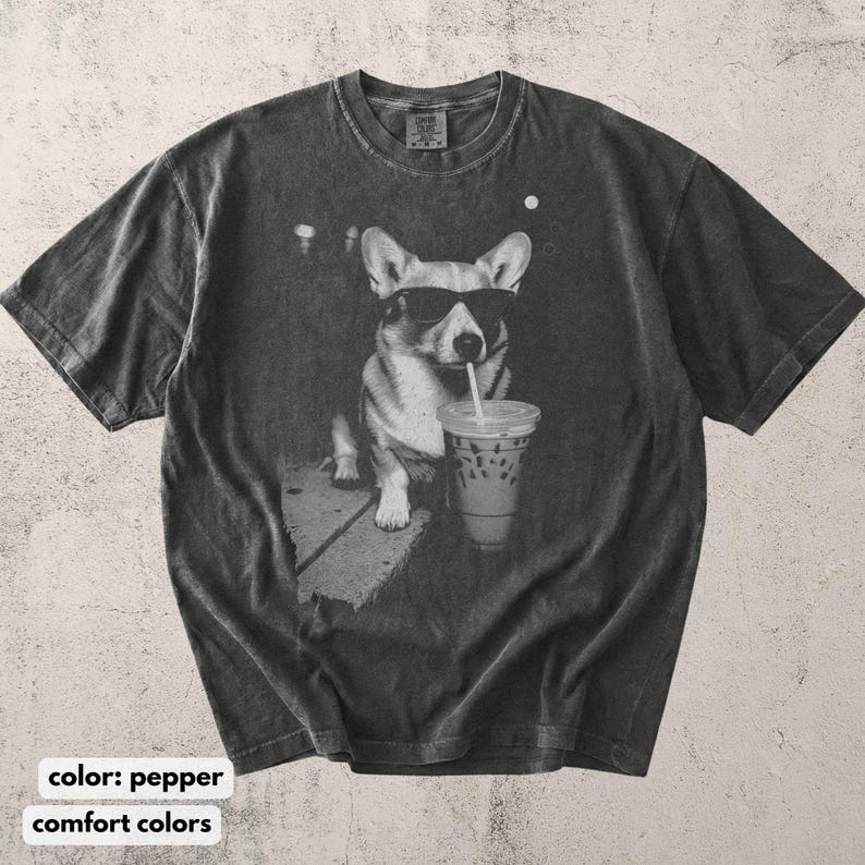 May include: Pepper-colored t-shirt featuring a graphic of a corgi wearing sunglasses and drinking from a to-go cup with a straw. The dog is sitting on a sidewalk. The shirt has a vintage, worn look.