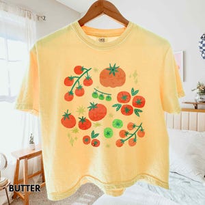 Puede incluir: Una camiseta amarilla con un dise&ntilde;o de tomate. El gr&aacute;fico presenta tomates rojos, hojas verdes y estrellas verdes. La palabra "BUTTER" est&aacute; impresa en la parte inferior izquierda de la imagen.