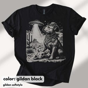 以下が含まれることがあります： 黒いTシャツ。カウボーイハットとバンダナを身に着けた犬がUFOを見上げているグラフィック。デザインには、頭蓋骨とサボテンのある砂漠の風景が含まれています。「color: gildan black」と「gildan softstyle」の文字が下にあります。