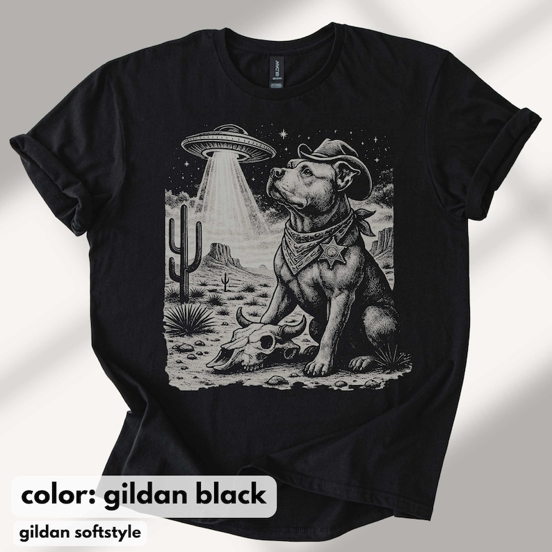 以下が含まれることがあります： 黒いTシャツ。カウボーイハットとバンダナを身に着けた犬がUFOを見上げているグラフィック。デザインには、頭蓋骨とサボテンのある砂漠の風景が含まれています。「color: gildan black」と「gildan softstyle」の文字が下にあります。