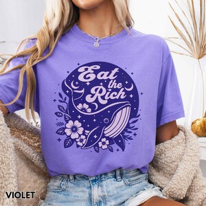 May include: Purple t-shirt featuring a whale graphic with the text "Eat the Rich" in a retro font. The whale is surrounded by stars and flowers.