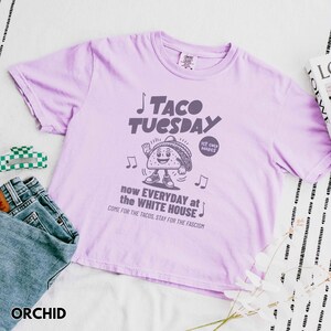 Pode incluir: Camiseta cor de orqu&iacute;dea com a inscri&ccedil;&atilde;o "TACO TUESDAY" e um personagem de taco de desenho animado. A camiseta tamb&eacute;m apresenta as frases "now EVERYDAY at the WHITE HOUSE" e "COME FOR THE TACOS, STAY FOR THE FASCISM".