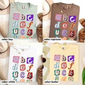 May include: Four t-shirts in bay, espresso, white, and butter colours, each with a colourful graphic of the letters "abc def UCK". The shirts are displayed with accessories like sandals and necklaces.