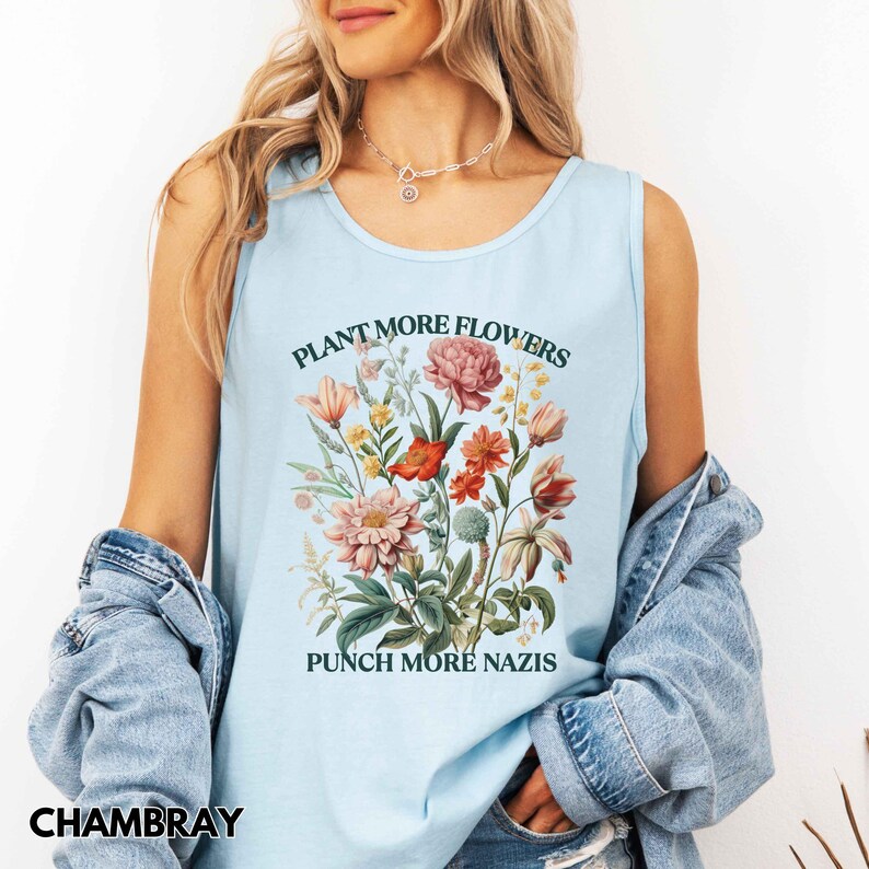 Op de afbeelding: Lichtblauwe tanktop met de tekst "PLANT MORE FLOWERS" boven een bloemenillustratie en "PUNCH MORE NAZIS" eronder. De tanktop is gemaakt van een zacht materiaal.