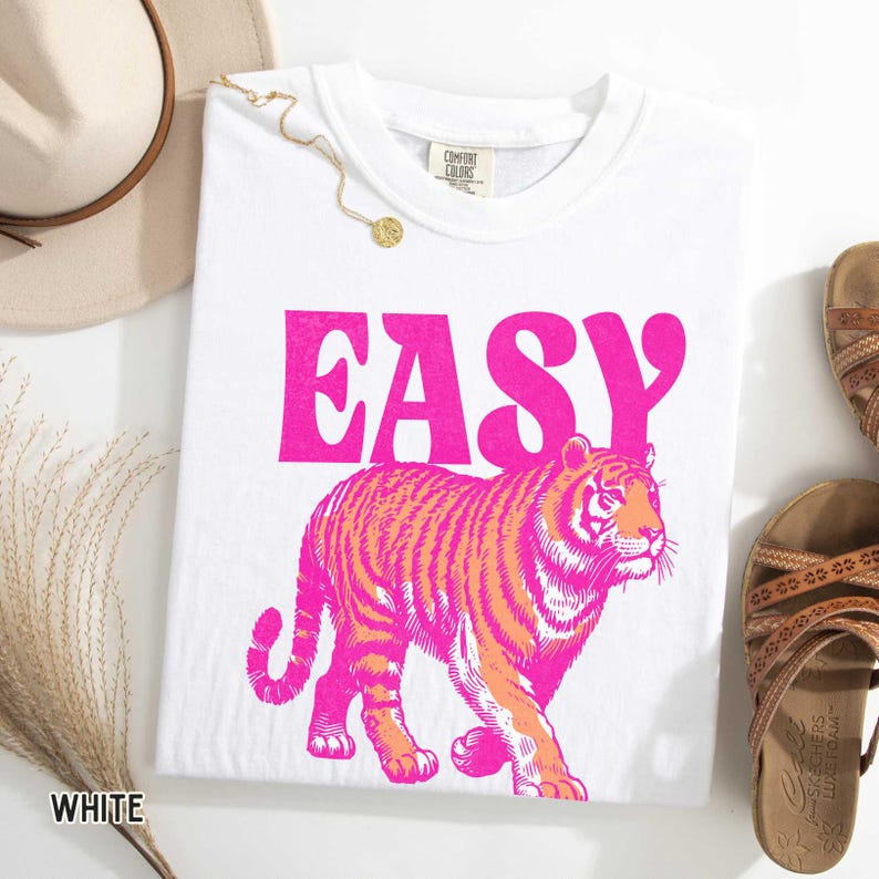 K&ouml;nnte beinhalten: Wei&szlig;es T-Shirt mit dem Wort "EASY" in leuchtend rosa Blockbuchstaben &uuml;ber einer rosa-orangenen Tigergrafik. Das Shirt liegt neben einem beigen Hut, einer goldenen Halskette und braunen Sandalen.