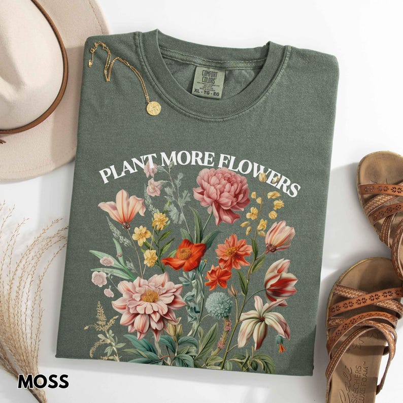 Op de afbeelding: Een mosgroen t-shirt met de witte tekst "PLANT MORE FLOWERS". Het shirt heeft een bloemenontwerp met roze, rode en gele bloemen. Een gouden ketting en een beige hoed zijn ook zichtbaar.