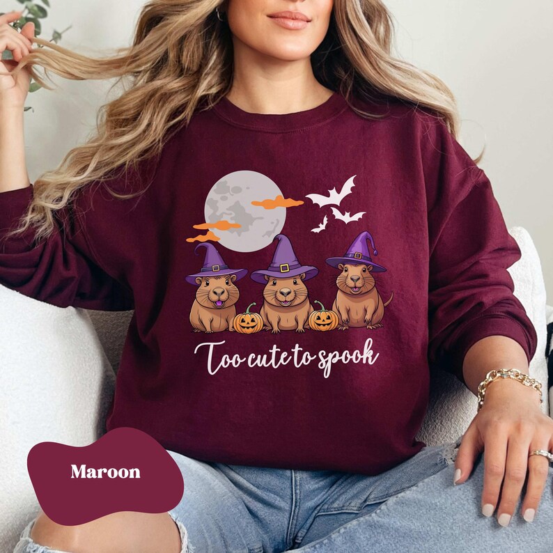 Op de afbeelding: Een maroon sweatshirt met een afbeelding van drie cartoon cavia's die toverhoeden dragen en pompoenen vasthouden. De tekst "Too cute to spook" is onder de cavia's gedrukt.
