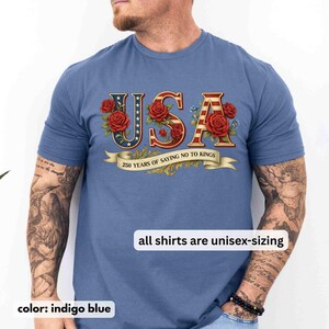 以下が含まれることがあります： インディゴブルーのTシャツ。赤、白、青で「USA」の文字が描かれ、赤いバラと青い花で飾られています。「250 YEARS OF SAYING NO TO KINGS」と書かれたバナーがあります。「all shirts are unisex-sizing」の文字も表示されています。