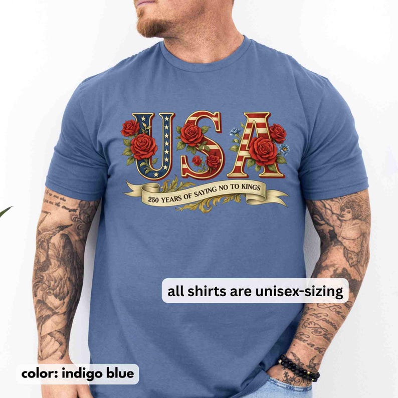 以下が含まれることがあります： インディゴブルーのTシャツ。赤、白、青で「USA」の文字が描かれ、赤いバラと青い花で飾られています。「250 YEARS OF SAYING NO TO KINGS」と書かれたバナーがあります。「all shirts are unisex-sizing」の文字も表示されています。