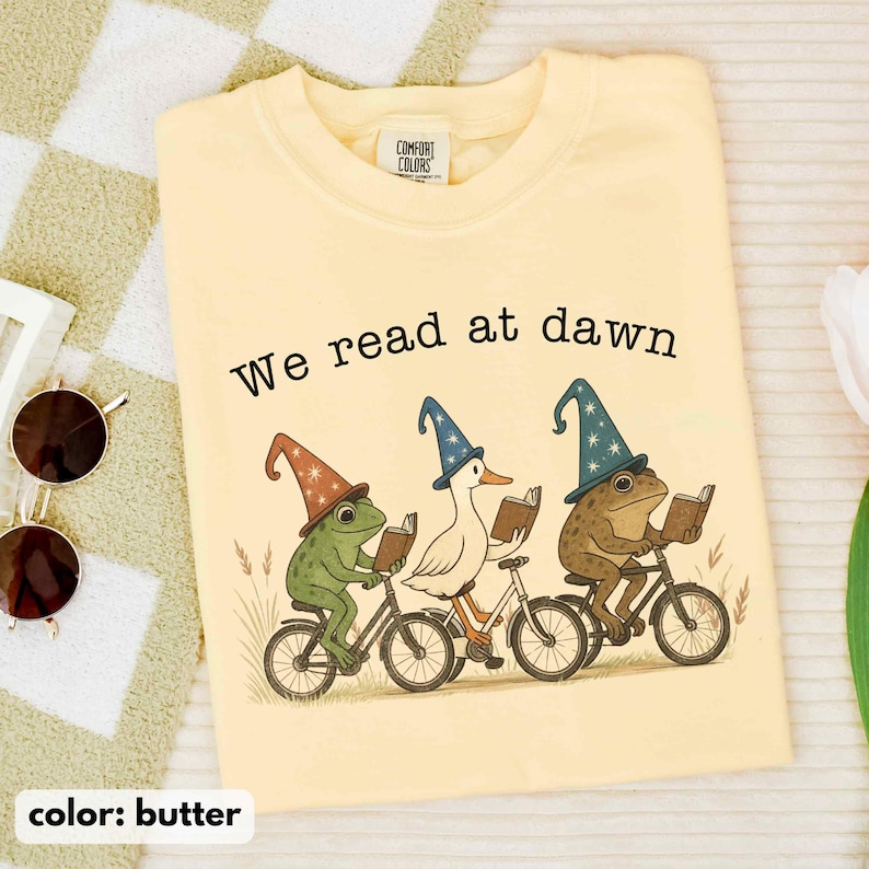 May include: Butter-colored Comfort Colors t-shirt with the text "We read at dawn." The graphic features a frog, a duck, and a frog riding bicycles and reading books, wearing wizard hats. The shirt is a light yellow color.