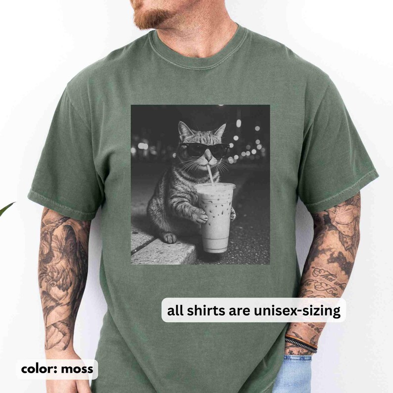 May include: Moss green unisex t-shirt featuring a black and white image of a cat wearing sunglasses and drinking an iced coffee. The text "all shirts are unisex-sizing" is printed below the image. The shirt is a solid color.