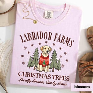 K&ouml;nnte beinhalten: Ein pinkfarbenes T-Shirt mit einem wei&szlig;en Labrador Retriever, der einen roten Pullover tr&auml;gt, vor zwei gr&uuml;nen Weihnachtsb&auml;umen. Der Text "Labrador Farms Christmas Trees Locally Grown, Cut by Paw" ist auf dem T-Shirt aufgedruckt.