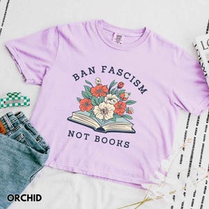 Op de afbeelding: Lila T-shirt met de tekst "BAN FASCISM NOT BOOKS" boven een bloemenillustratie. Het shirt heeft korte mouwen en een ronde hals. Een spijkerbroek en een groen-witte haarclip zijn zichtbaar.