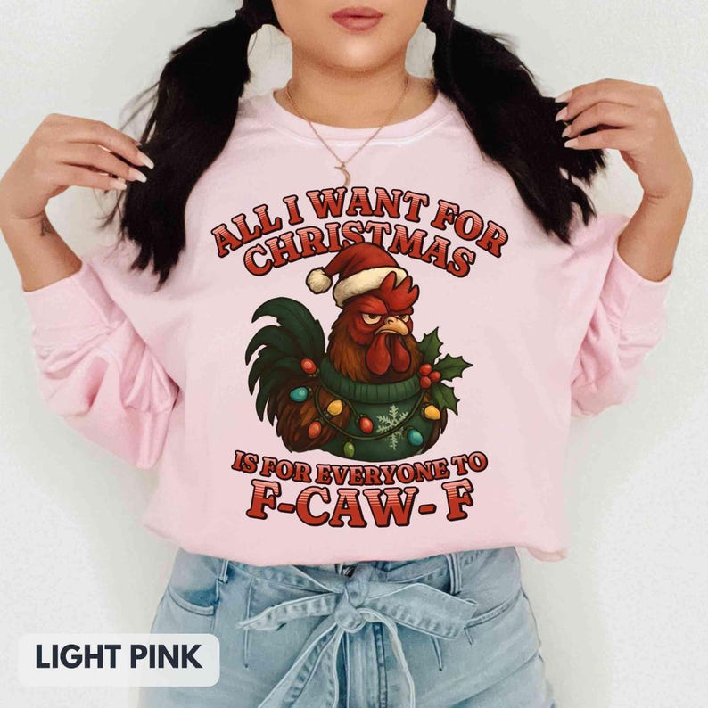 May include: Light pink sweatshirt with a Christmas rooster graphic. The rooster wears a Santa hat and a green sweater with Christmas lights. Text reads "ALL I WANT FOR CHRISTMAS IS FOR EVERYONE TO F-CAW-F."