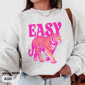 May include: An ash gray crewneck sweatshirt featuring the word EASY in bold pink letters above a graphic of a walking tiger rendered in vibrant pink and orange tones. The garment has long sleeves and a ribbed neckline, showcasing a casual design.