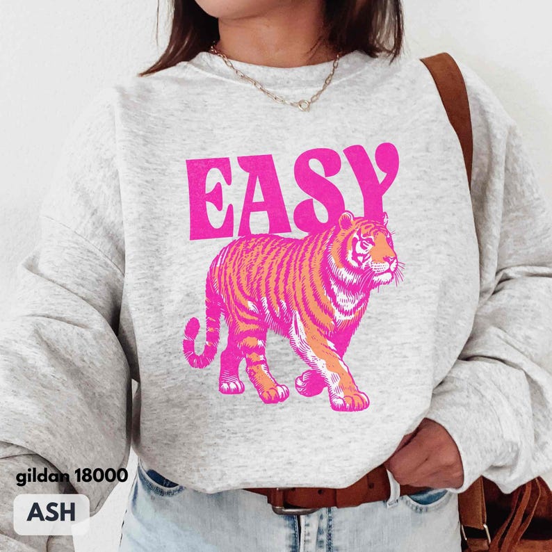 May include: An ash gray crewneck sweatshirt featuring the word EASY in bold pink letters above a graphic of a walking tiger rendered in vibrant pink and orange tones. The garment has long sleeves and a ribbed neckline, showcasing a casual design.