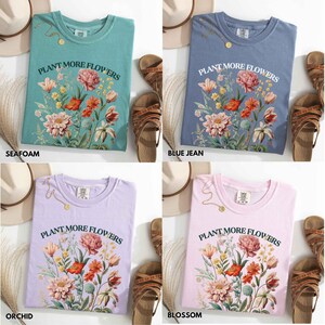 Op de afbeelding: Vier T-shirts in de kleuren zeegroen, spijkerbroekblauw, orchidee en bloesem. Elk shirt heeft de tekst "PLANT MORE FLOWERS" boven een bloemenontwerp met rode, roze en witte bloemen. Accessoires zijn sandalen en een hoed.
