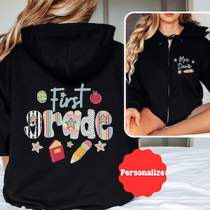 May include: Black zip-up hoodie with "First Grade" graphic on the back, featuring colorful letters, stars, a pencil, and a book. The front of the hoodie has the text "Mrs. Davis" with a pencil and stars. A red "Personalize" sticker is also visible.