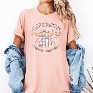 May include: A light pink t-shirt with the text "LAST CHAPTER 2025 before The Chapel AMANDA'S BACHELORETTE" in a floral design. The design includes books, flowers, and stars. The shirt has rolled-up sleeves.