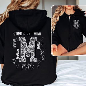 May include: Black zip-up hoodie with a white floral "M" design and the word "Mimi" in various placements. The hoodie has a hood and a front pocket. The design is repeated on the front of the hoodie.