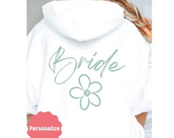 May include: White hoodie with the word "Bride" in a script font and a flower graphic in a light green color on the back. The hoodie has a drawstring hood and a kangaroo pocket. The word "Personalize" is on a pink sticker.