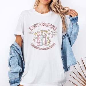May include: White t-shirt with a colorful graphic design. The design includes the text "LAST CHAPTER 2025 before the Chapel" and "AMANDA'S BACHELORETTE" in a whimsical font, along with floral and book illustrations.