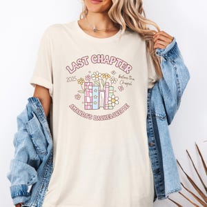 May include: Cream-colored t-shirt with a floral and book graphic. The graphic includes the text "LAST CHAPTER 2025 before the Chapel" and "AMANDA'S BACHELORETTE" in a playful font. The design features flowers and books.