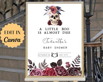 May include: A white sign with a skull wearing a floral crown and the text "A little boo is almost due Tashantha's Baby Shower October 29 ANY CITY". The sign is on a white easel.
