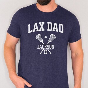 May include: Navy blue t-shirt with the text "LAX DAD" in white, with a lacrosse stick and ball graphic. The text "JACKSON 13" is below the graphic.