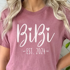 May include: A mauve t-shirt with white text that reads "Bibi -EST. 2024- " with two small hearts above the word "Bibi".